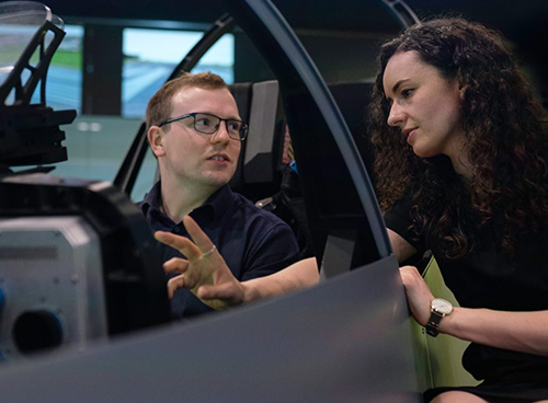 Man sitting in an airplane simulator, next to a woman gesturing directions